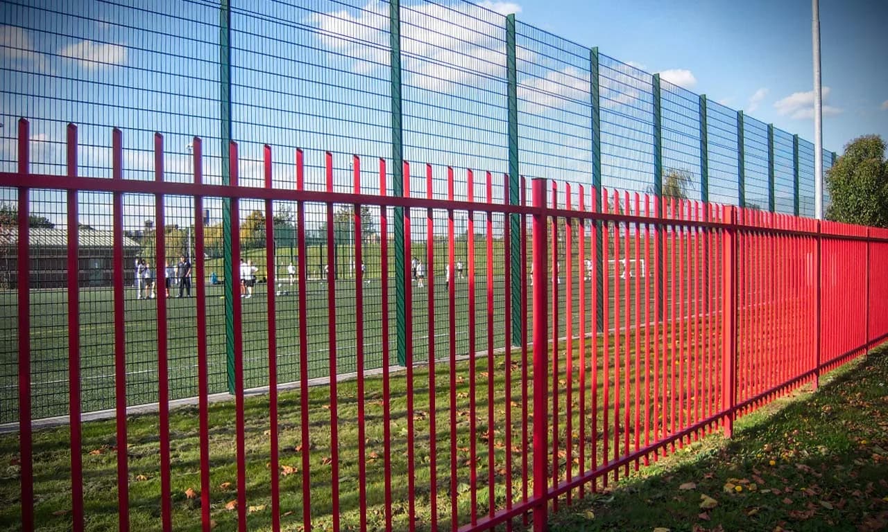 Red steel palisade school perimeter fence with green anti-climb mesh around a school sports field — Fenced.ca
